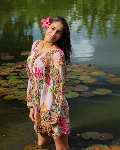 A Woman in Printed Dress Standing on the Lake with Pink Flower on Her Ear