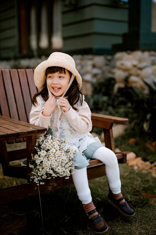 Full body of positive Asian girl wearing straw hat looking away while sitting on wooden chair with white flowers on terrace of building on blurred background