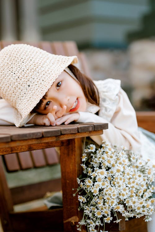 Cute Asian girl with head on wooden table wearing straw hat looking at camera while sitting on wooden chair with white flowers against blurred background