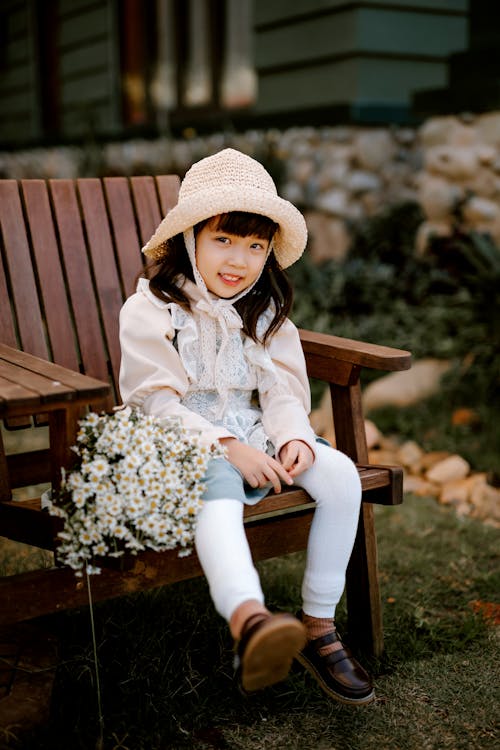 Full body of content Asian girl wearing straw hat looking at camera while sitting on wooden chair with bouquet of flowers in backyard on blurred background