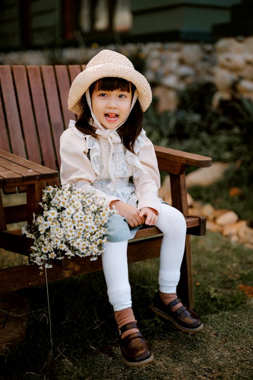 Full body of positive Asian kid with white flowers wearing straw hat looking away while sitting on wooden armchair in backyard on blurred background