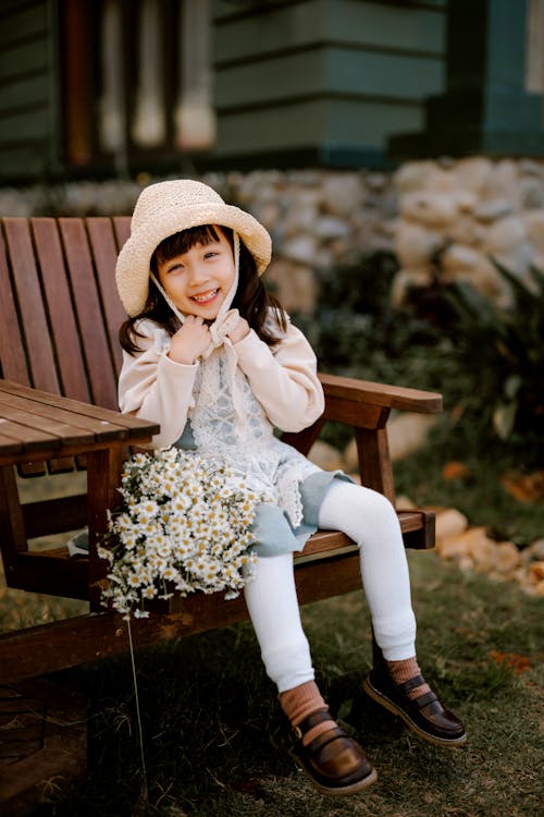 Full body of cheerful Asian kid looking at camera while sitting on wooden chair with white flowers in backyard on blurred background