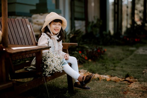 Full body of positive Asian kid wearing straw hat looking away while sitting on wooden bench with bouquet of white flowers on blurred terrace