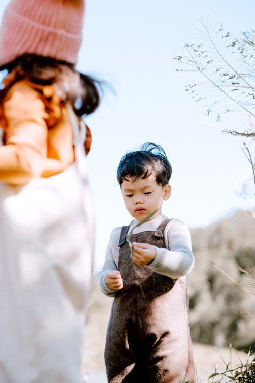 From below of cute Asian kid with leaf in hand standing in countryside near unrecognizable girl in hat against blurred background