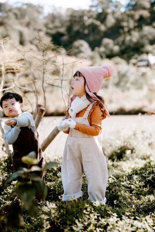 Free Full body of cheerful Asian boy and girl wearing overalls standing on grassy field against lush trees on blurred background Stock Photo