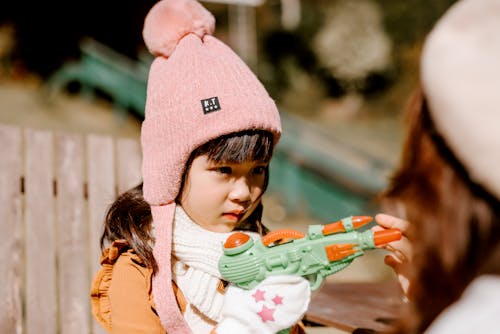 Concentrated Asian kid in hat with toy pistol looking at anonymous woman while standing near wooden fence on blurred background