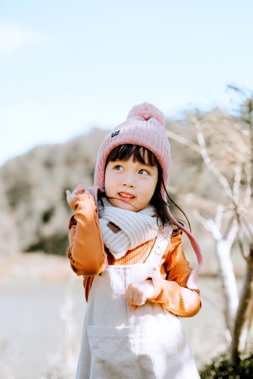 Cheerful Asian kid wearing hat looking up with arm bent in elbow while standing in sunny nature on blurred background