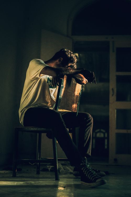 Free A man sits pensively on a chair in a dimly lit indoor setting, conveying a moody atmosphere. Stock Photo