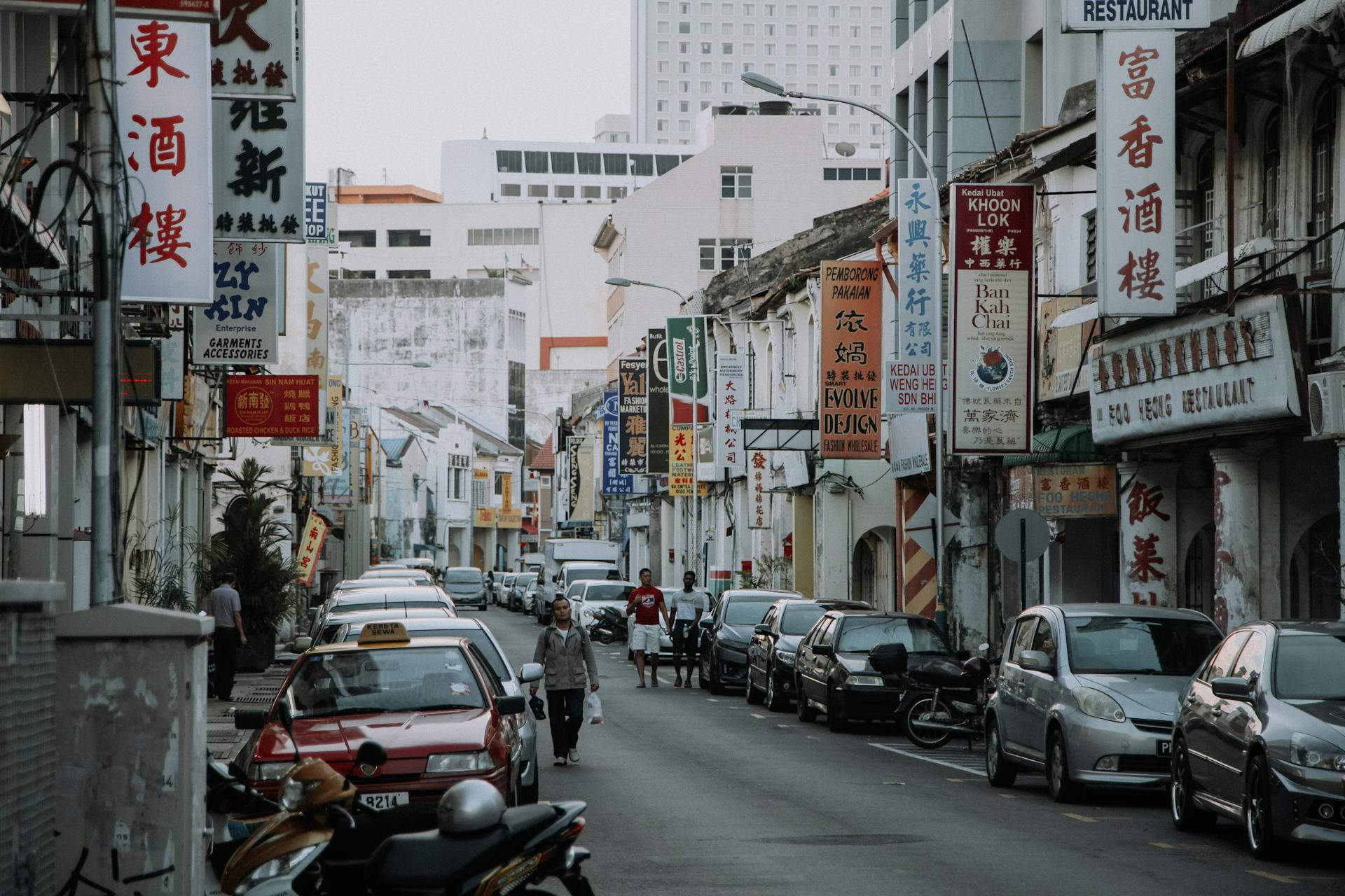 People walking on narrow asphalt roadway with parked cars between houses with hieroglyphs written on signboards on street in city