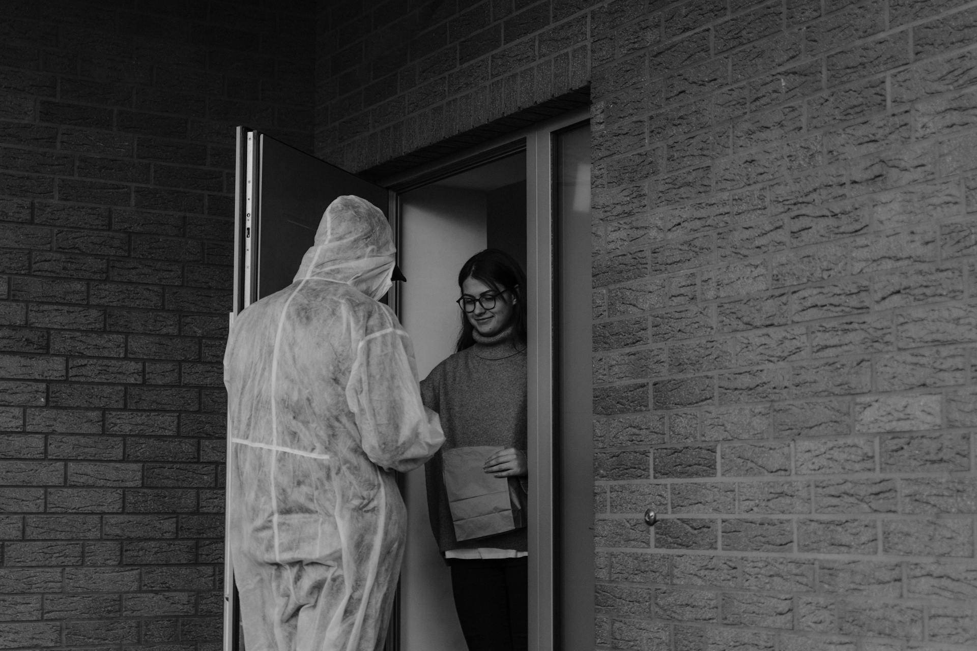 A delivery person in protective gear hands a package to a woman at the door in a contactless method.