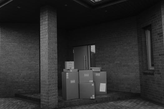 Grayscale photo showing stacked parcels by a building's doorway, evoking modern delivery themes.