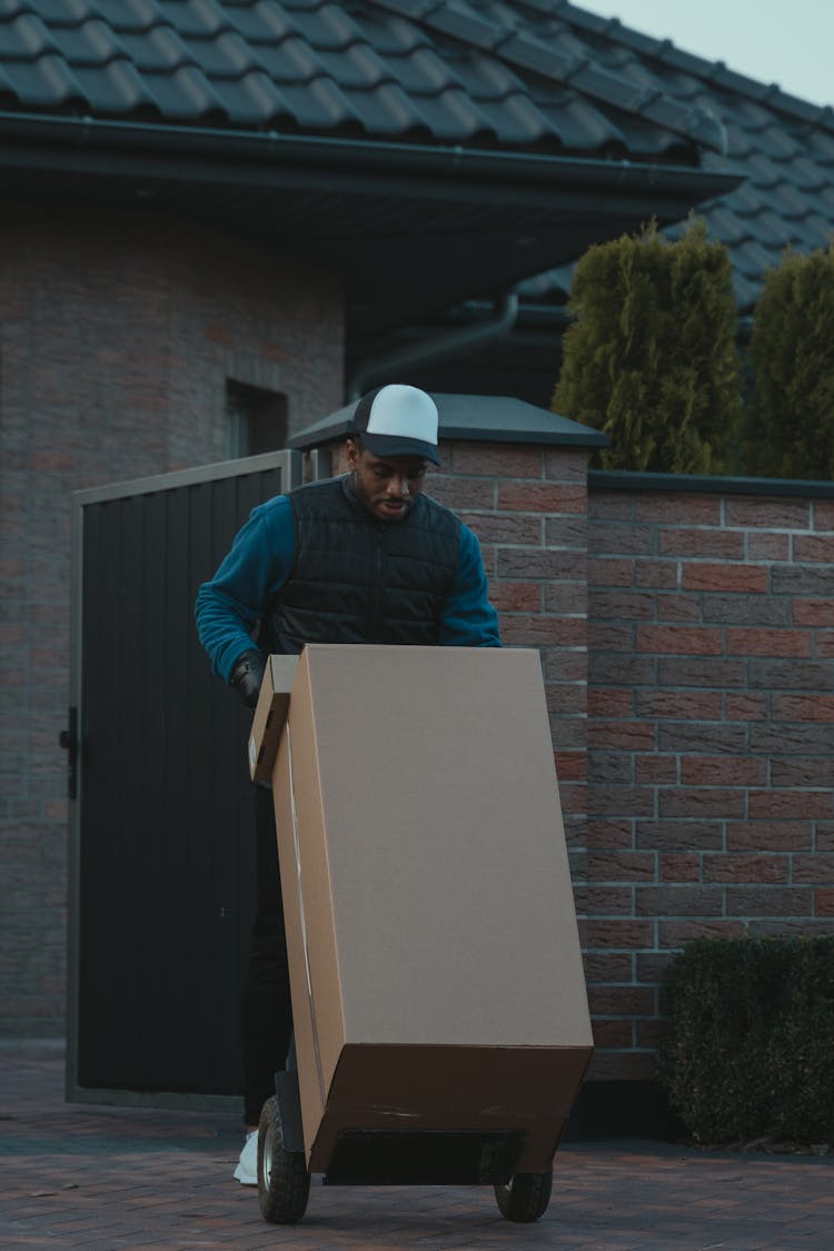 A Man In Black Puffer Vest Pushing A Trolley With Carton Boxes