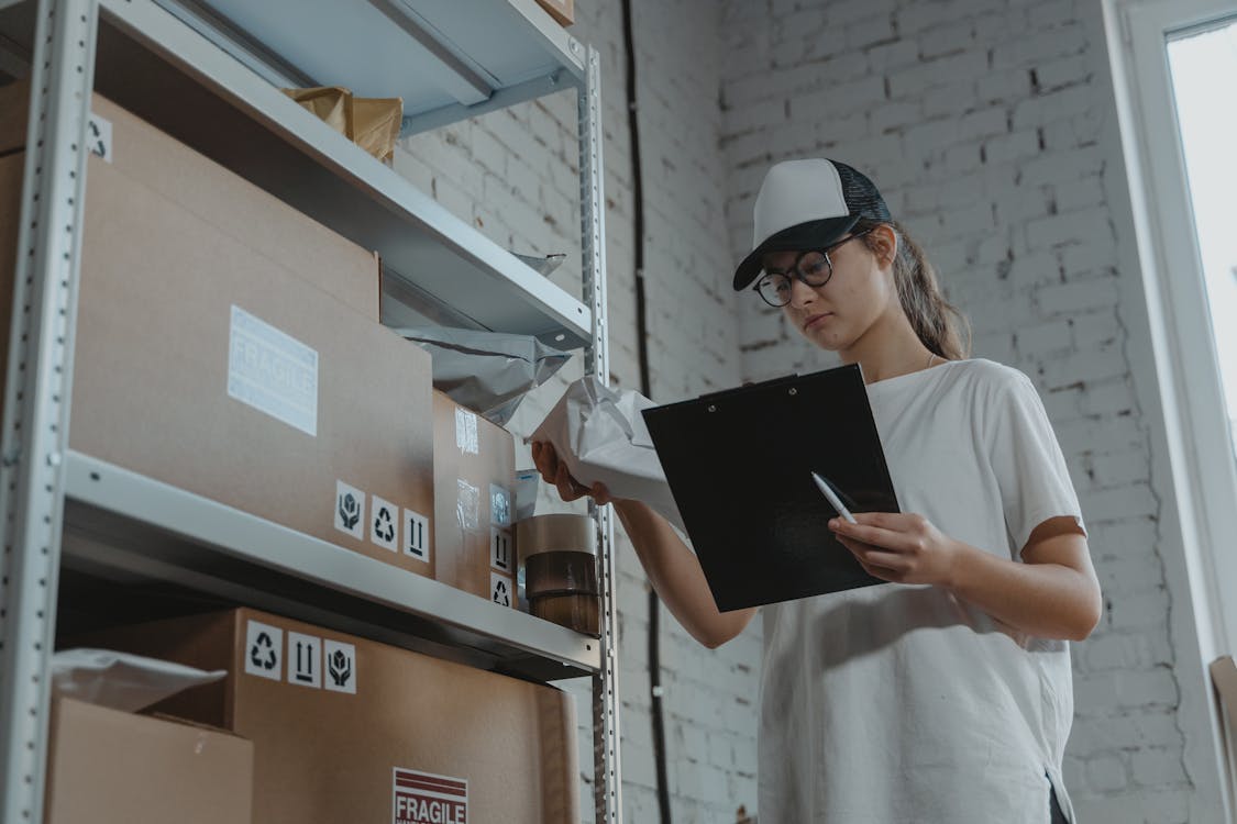 Free Warehouse worker organizing packages on shelf with clipboard. Stock Photo