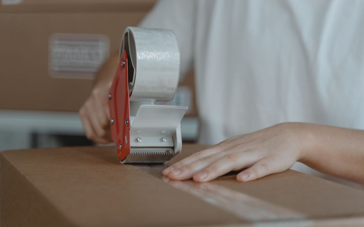 A person sealing a cardboard box with tape, emphasizing packaging and shipping processes.