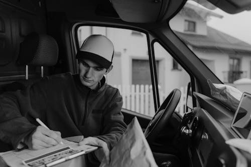 Grayscale Photo of Delivery Man writing on a Paper on Top of Carton 