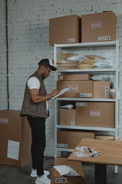 Free Business Man writing on a Paper while standing beside a Shelf of Boxes Stock Photo