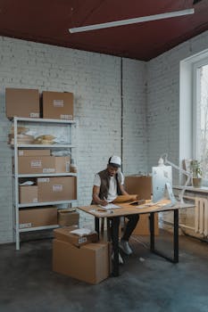 A young man in a cap organizing packages in a small warehouse office.