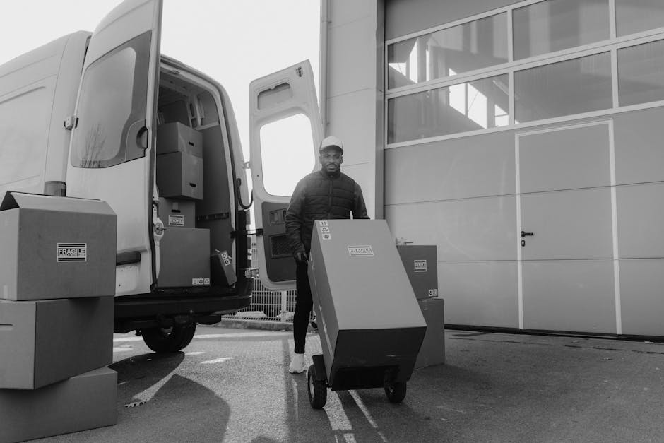Monochrome image of a delivery man with boxes on a trolley near a van. Snapped by Tima Miroshnichenko