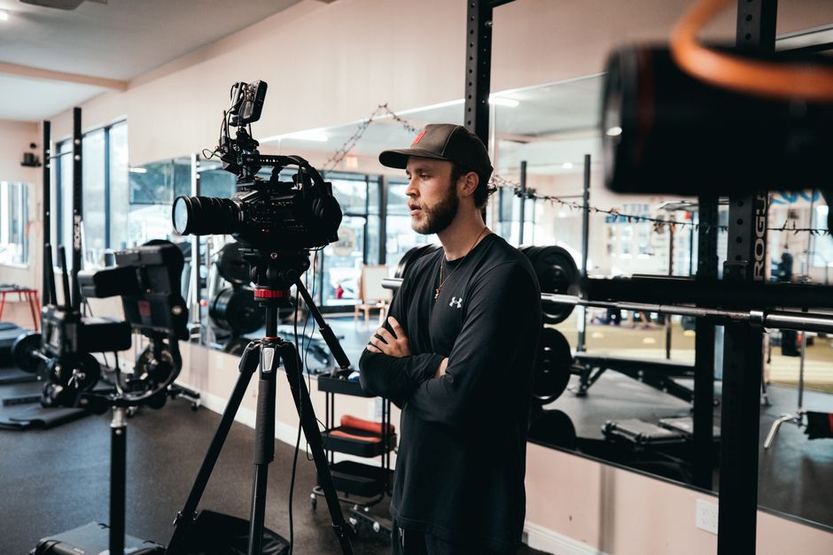 A cinematographer directs a workout video in a gym, using professional camera gear.