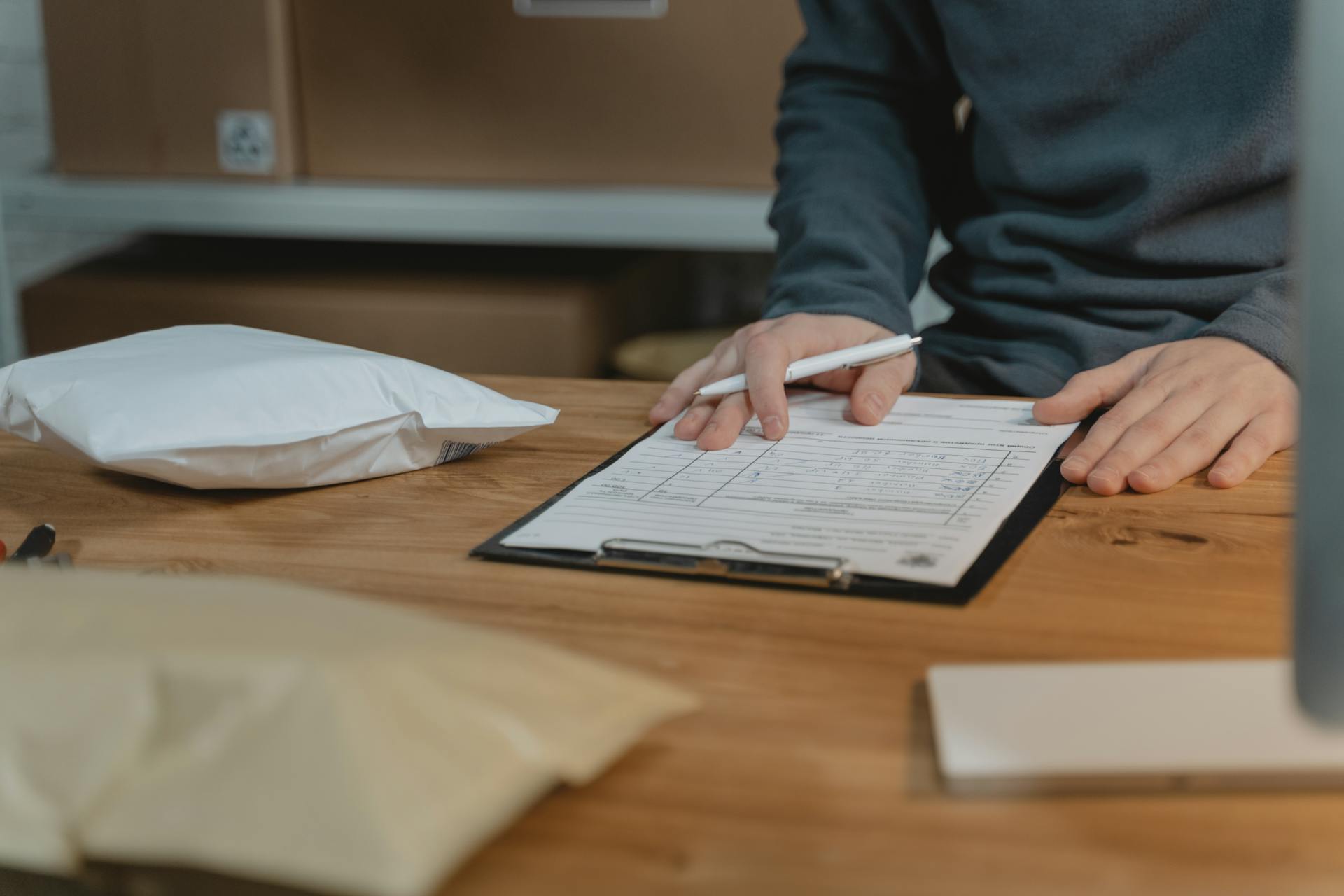 Close-up of a person filling out a form on a clipboard with packages nearby in an office setting.