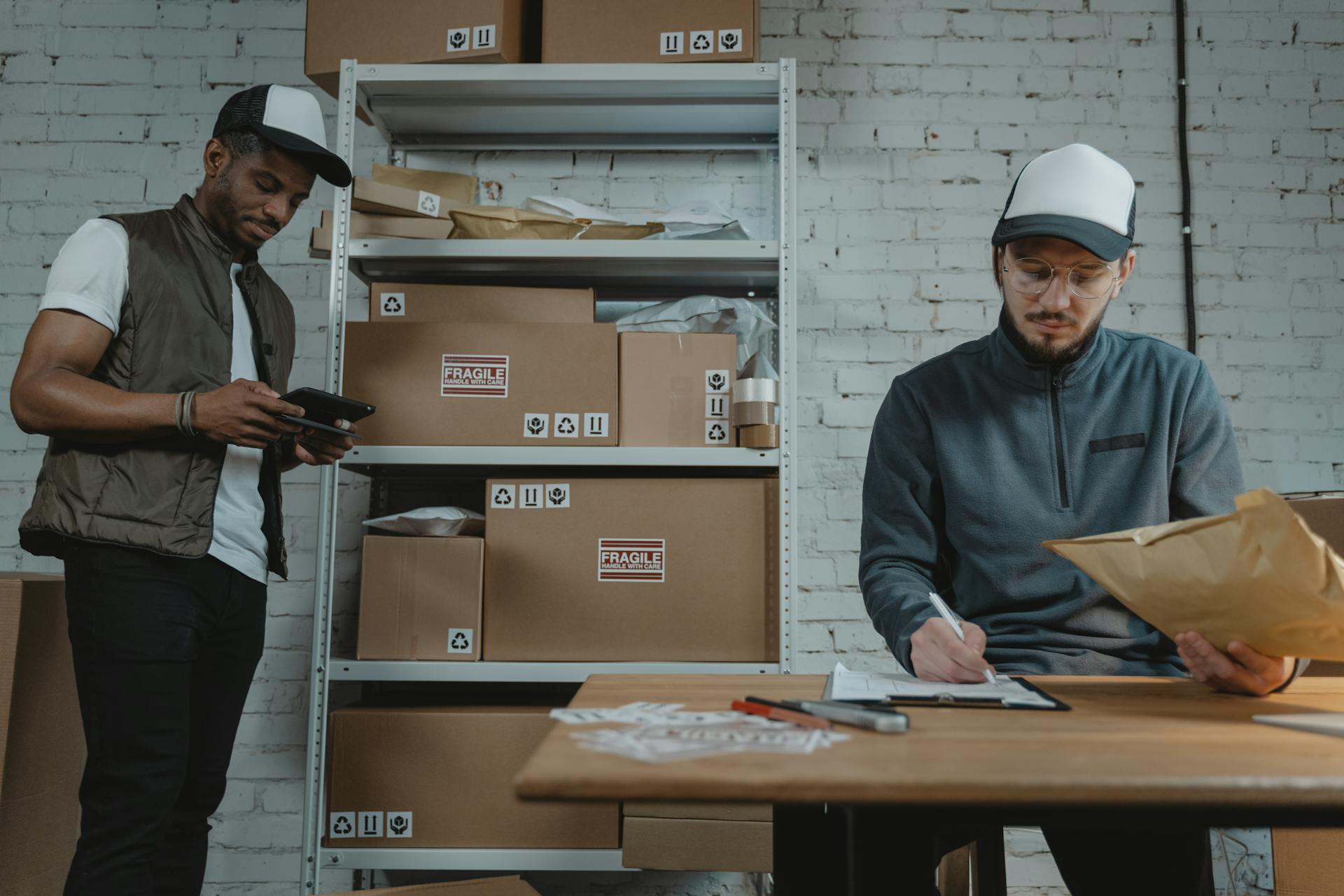 Warehouse employees organizing shipments with packages and documentation in a brick-walled workspace.