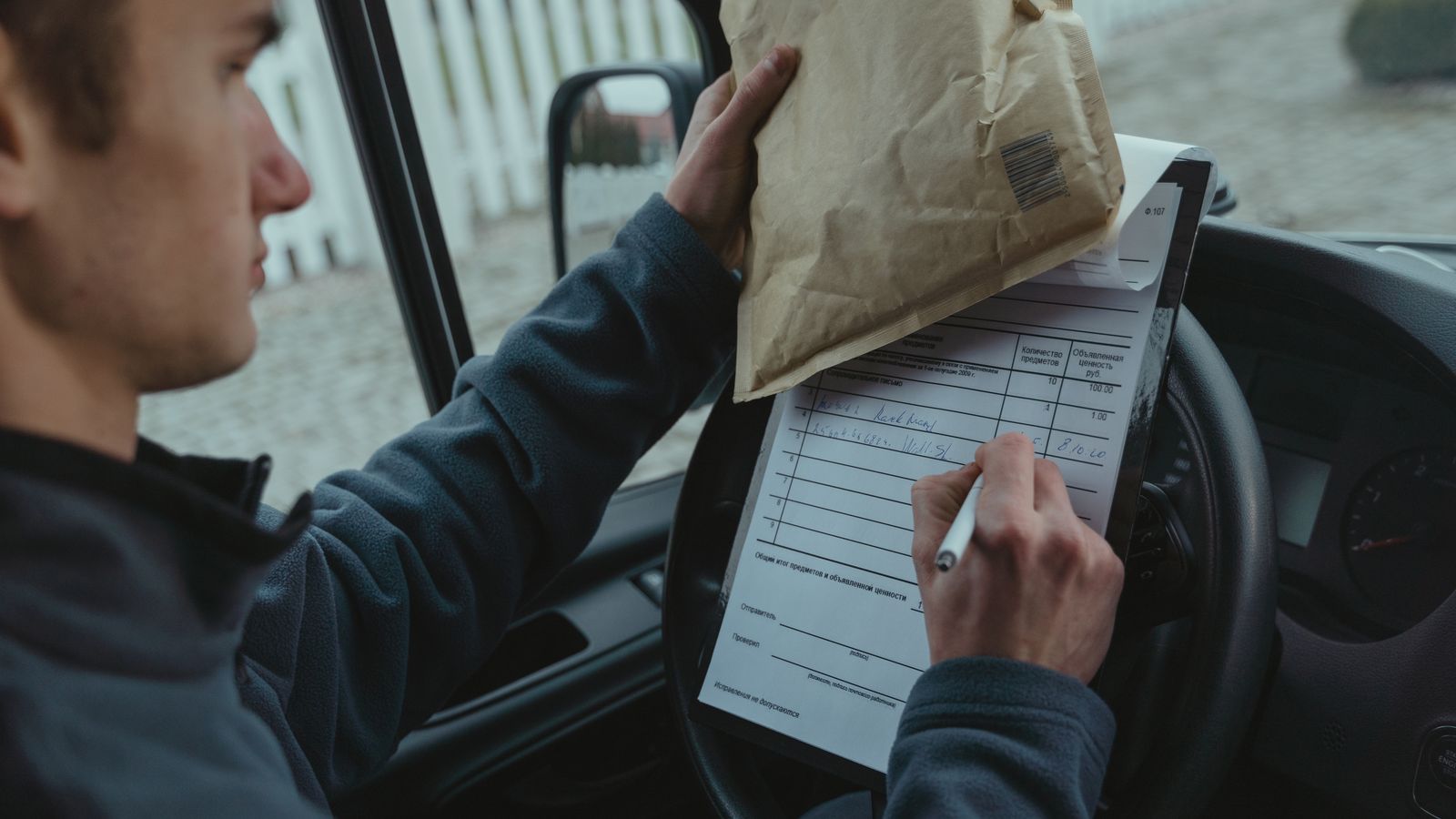Courier signs delivery documents inside van while holding package, representing logistics and efficient mail service.