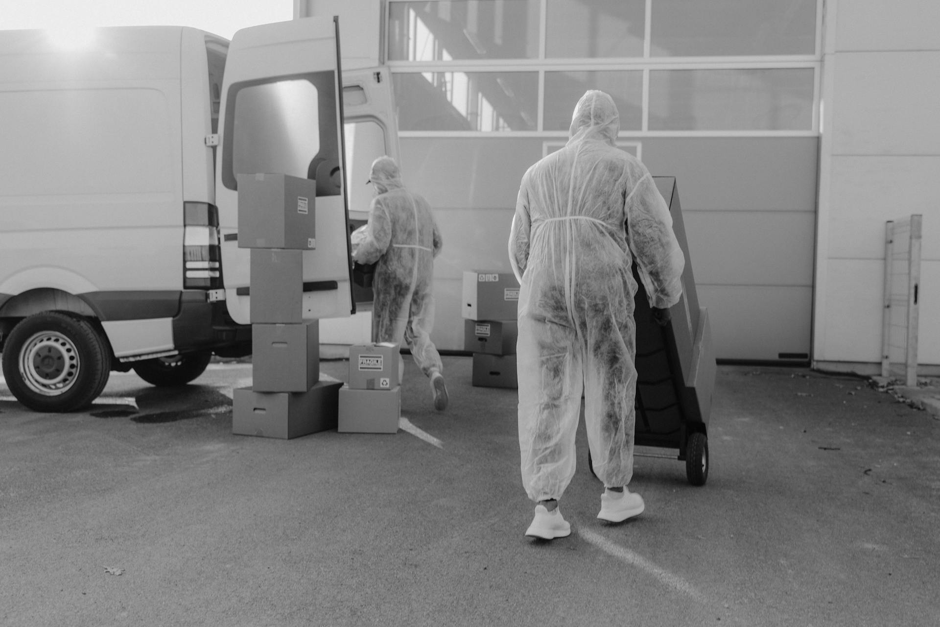 Workers in protective gear unload packages from a delivery van outside a warehouse.