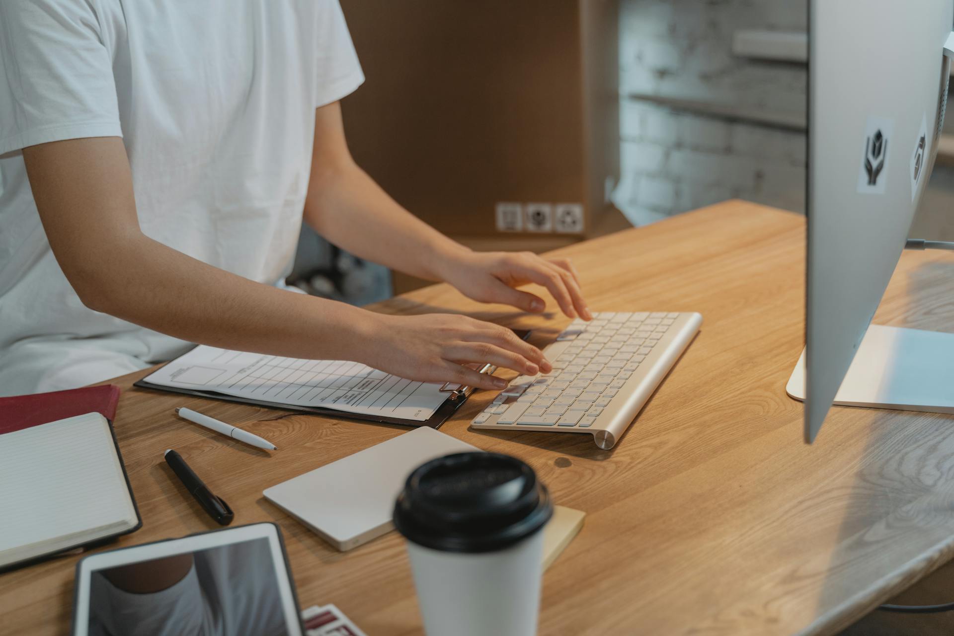 A person typing on a keyboard with documents on a wooden desk. Modern workspace setup.