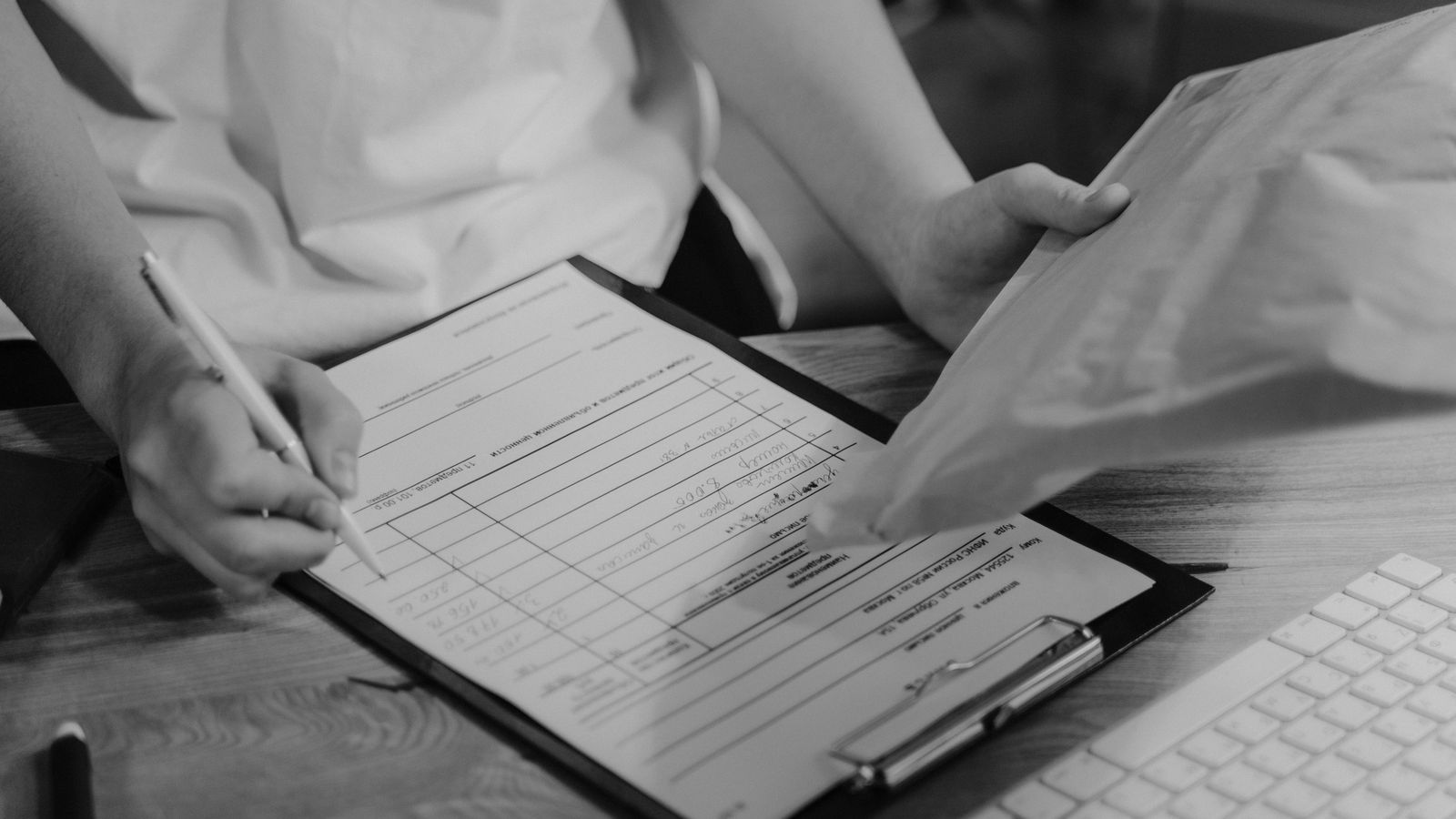 Black-and-white photo of a person writing on a clipboard with a keyboard visible.