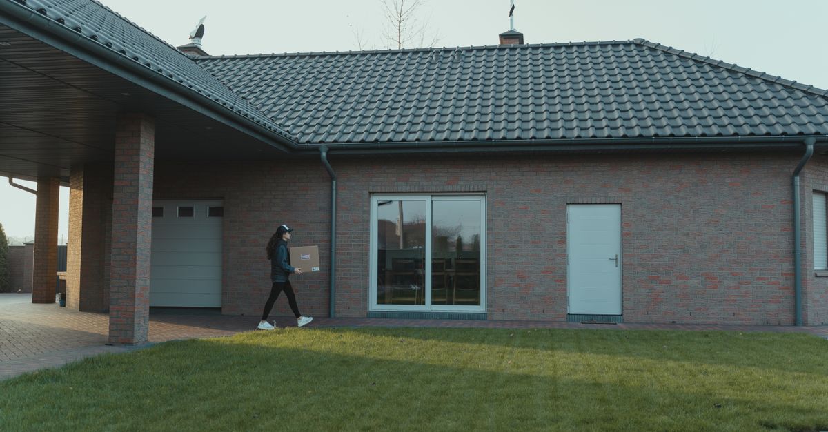 Person delivering package to a modern brick house with a tile roof and green lawn.