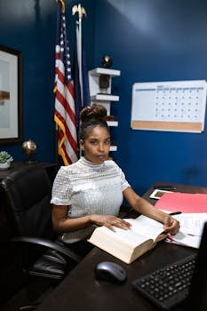 African American woman in office setting, reading a book at her desk.