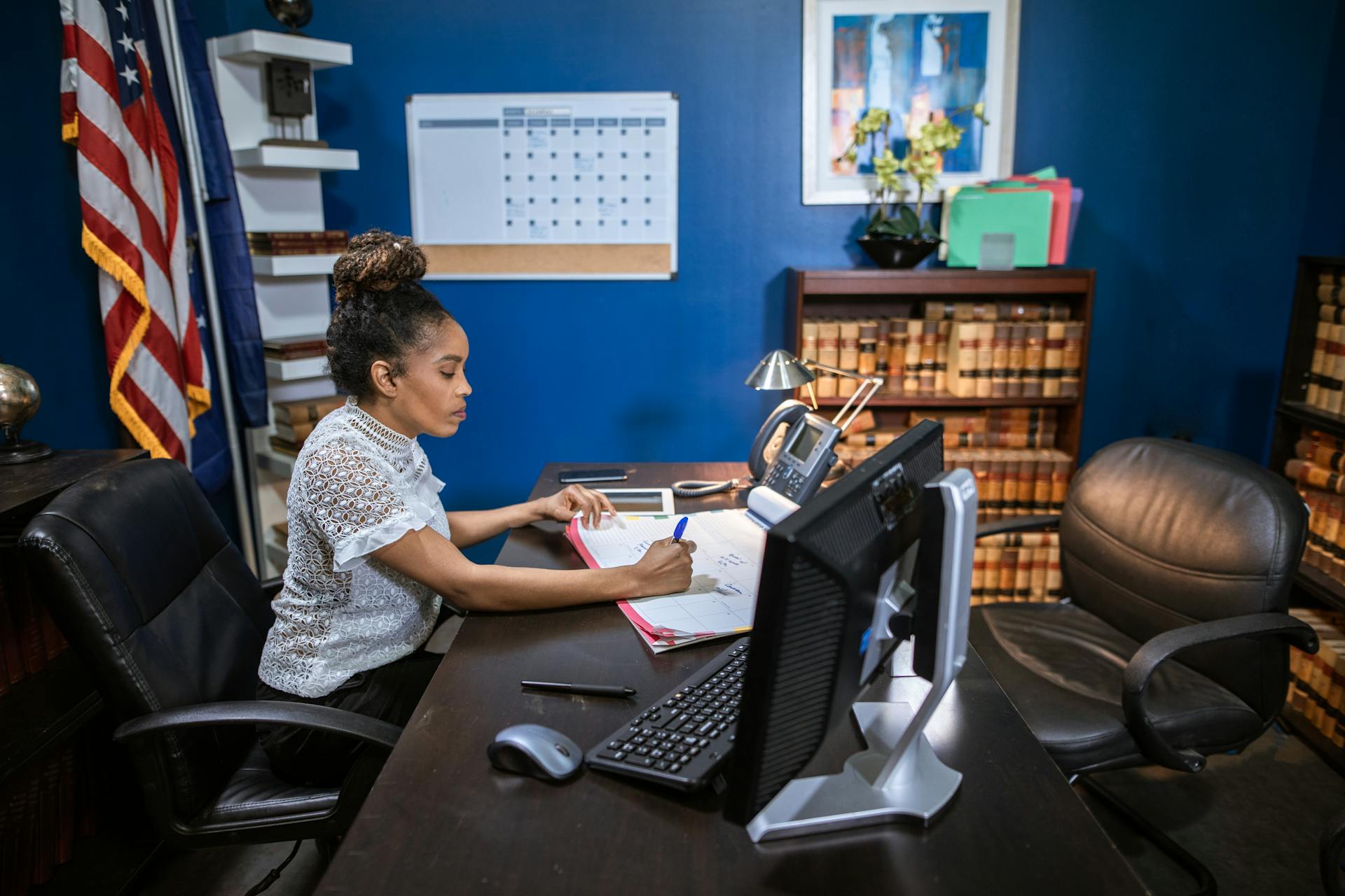 A woman writing at her desk in a professional office with legal books and an American flag.