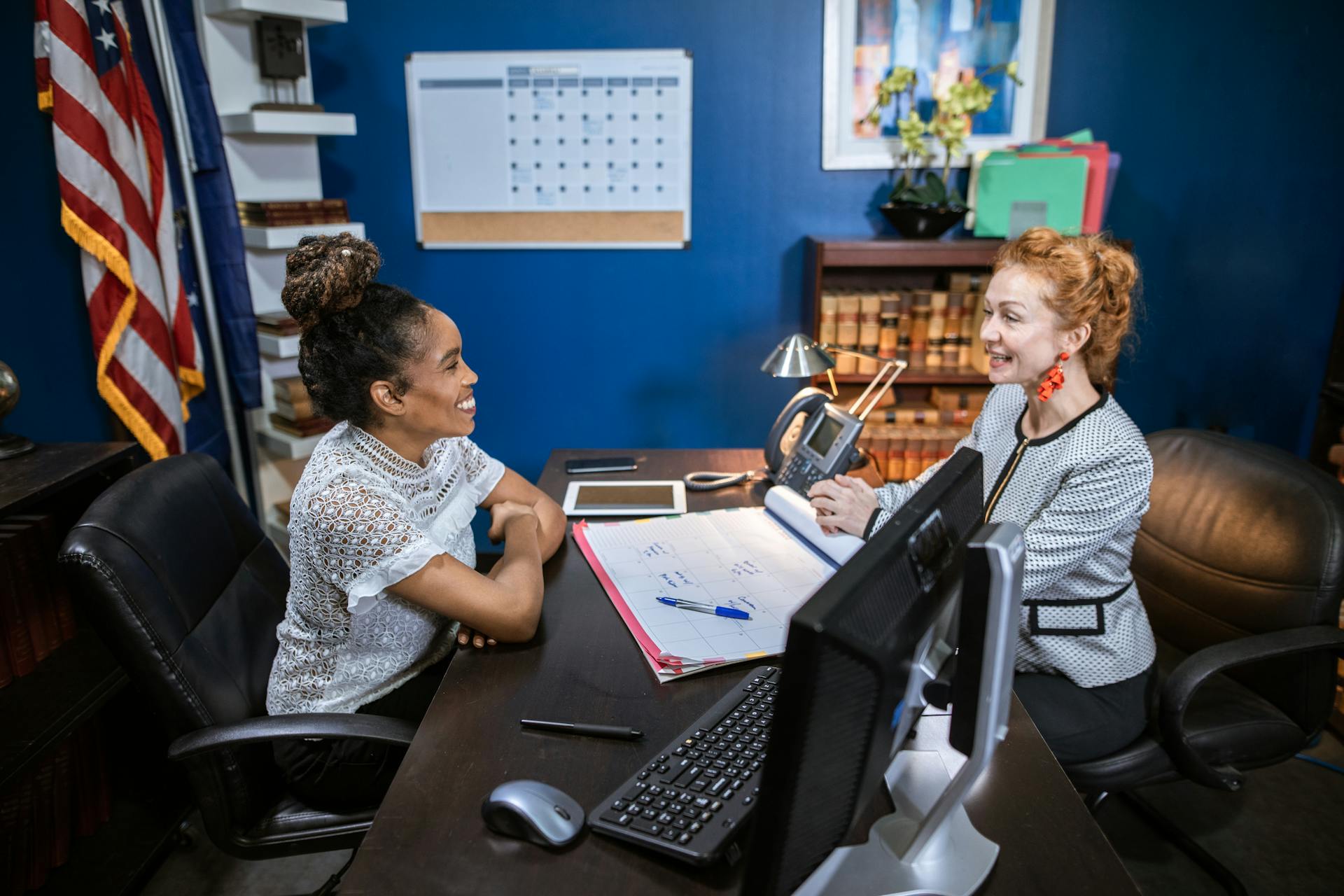 Two professional women discussing at an office desk, showcasing leadership and collaboration.