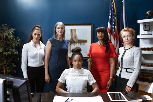 Group of Women Standing Beside Table with one woman seated in middle.