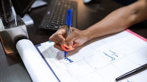 Detailed shot of a person's hand writing notes on a calendar using a blue marker.