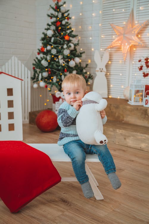 Adorable toddler in warm sweater sitting on bench with toy against Christmas tree and various glowing garlands