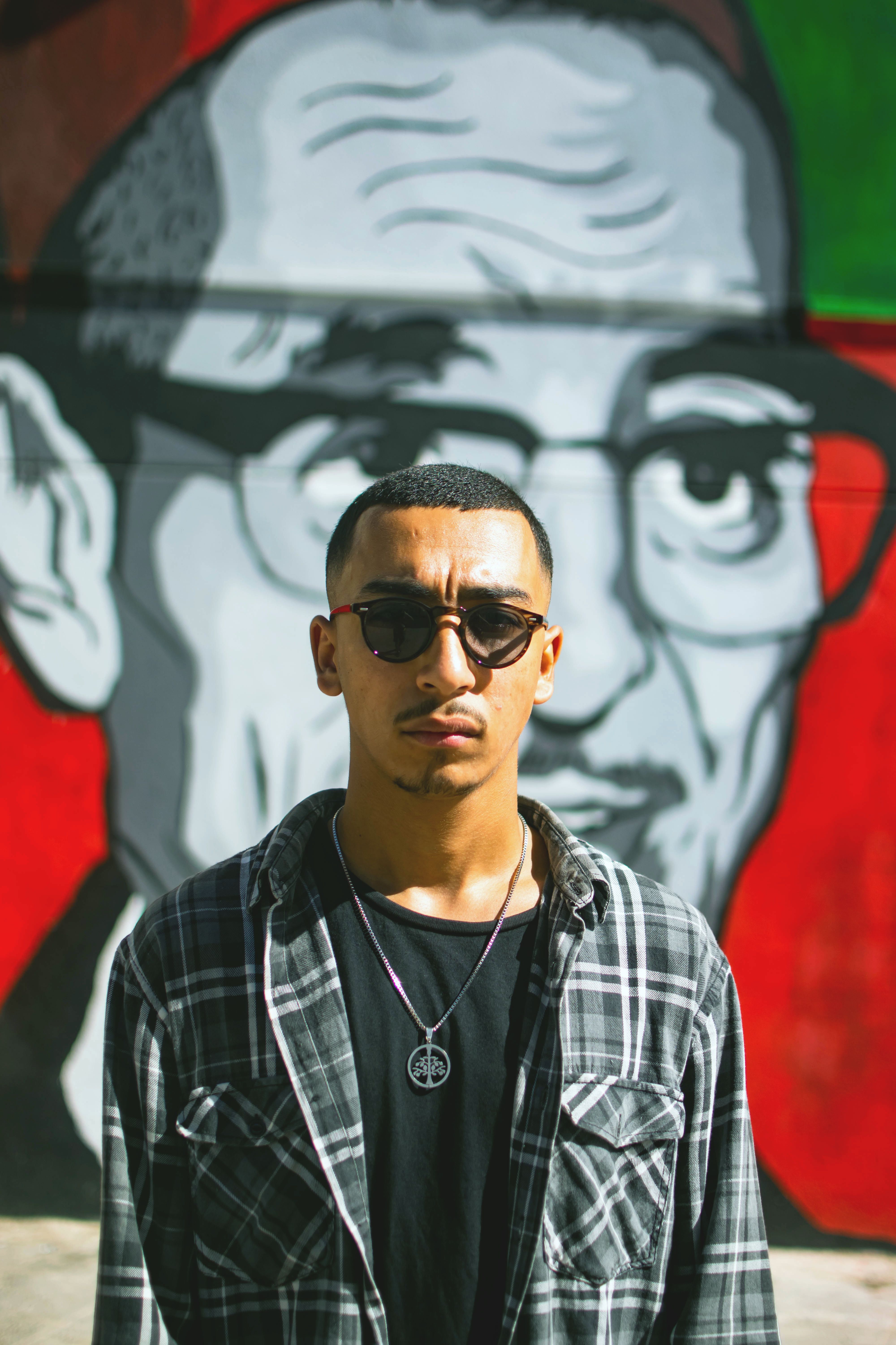 Cool urban man wearing sunglasses and plaid shirt standing against vibrant mural.