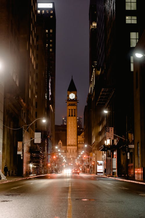 Free View of ancient tower clock through narrow illuminated street with road and tall residential buildings at dark time in city Stock Photo