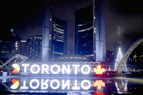Modern illuminated Toronto sign reflecting on ground located on street against contemporary skyscrapers at night time in city with glowing Christmas tree in distance