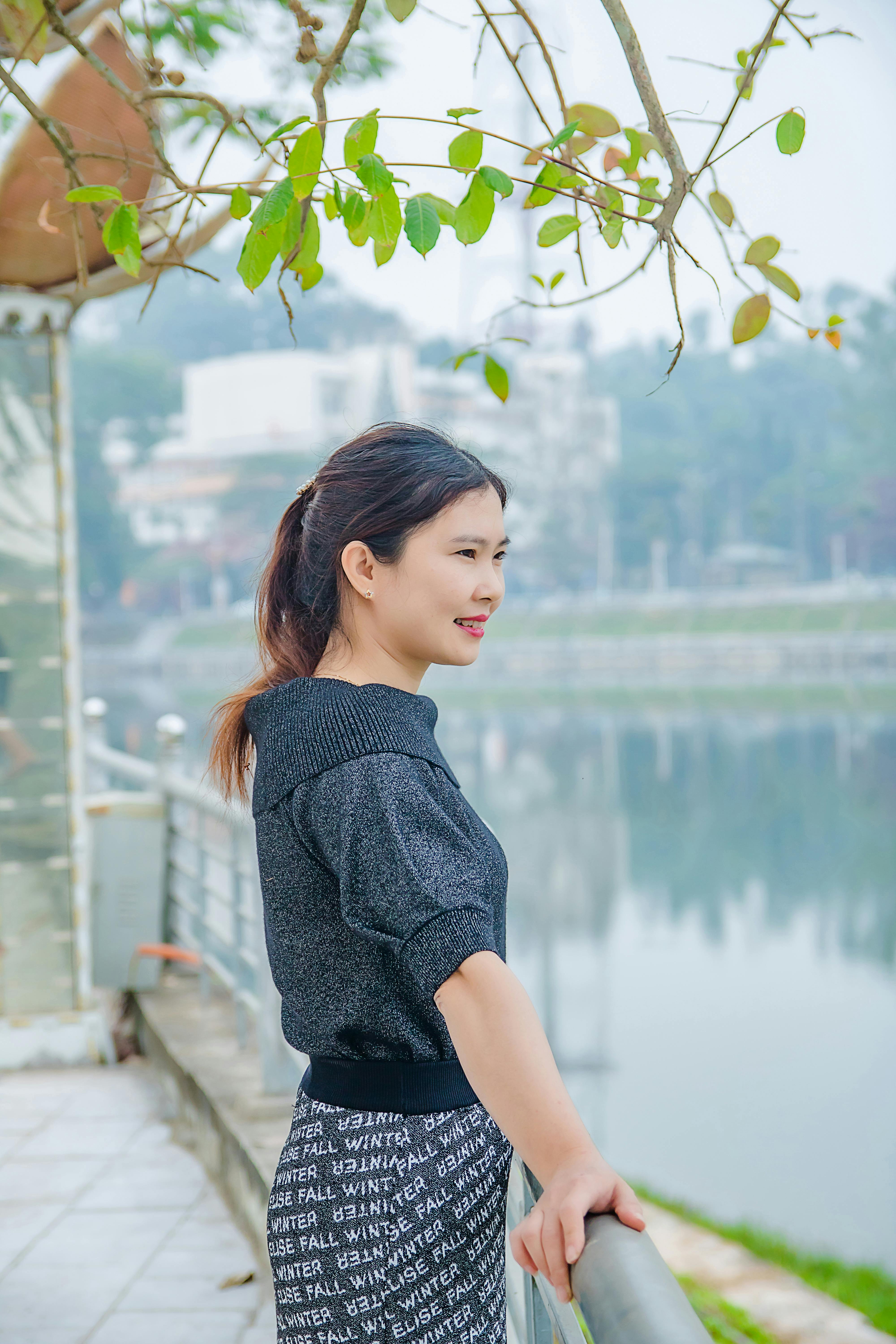 A young woman smiles as she stands by a lakeside railing in an urban setting.