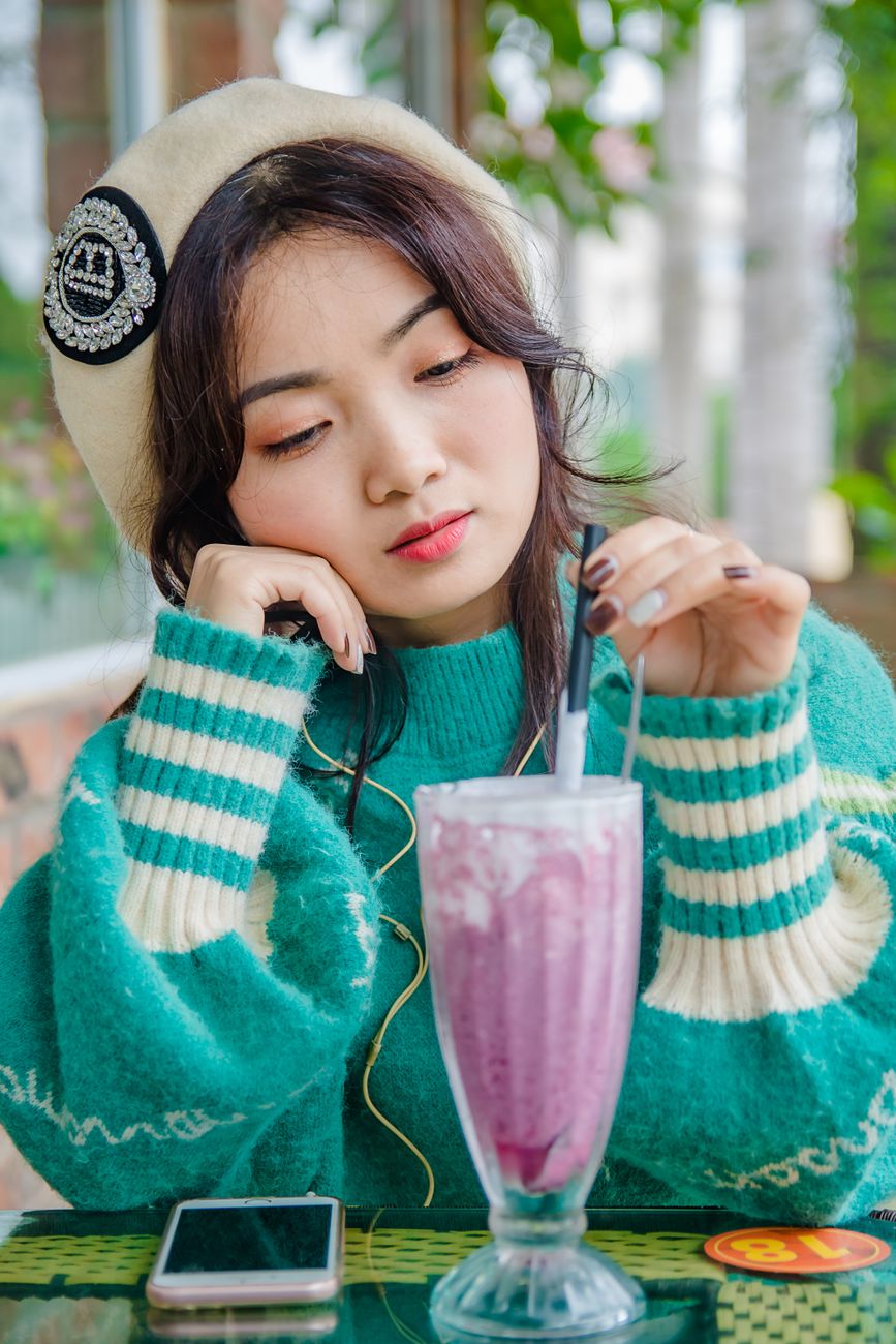Young woman in cozy sweater stirring a berry smoothie at a cafe, looking thoughtful.