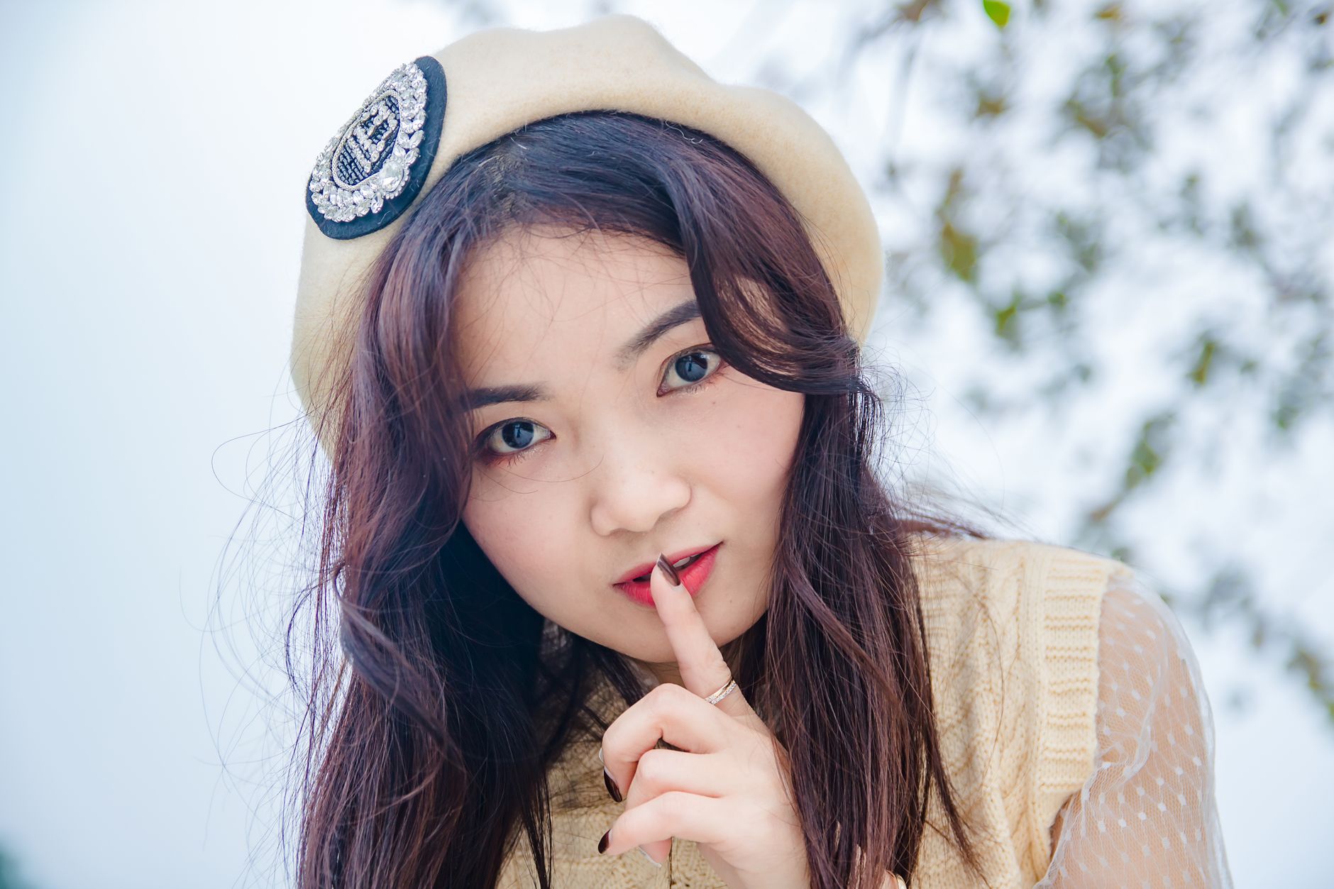 Close-up portrait of an Asian woman outdoors, signaling quiet with a finger to her lips.
