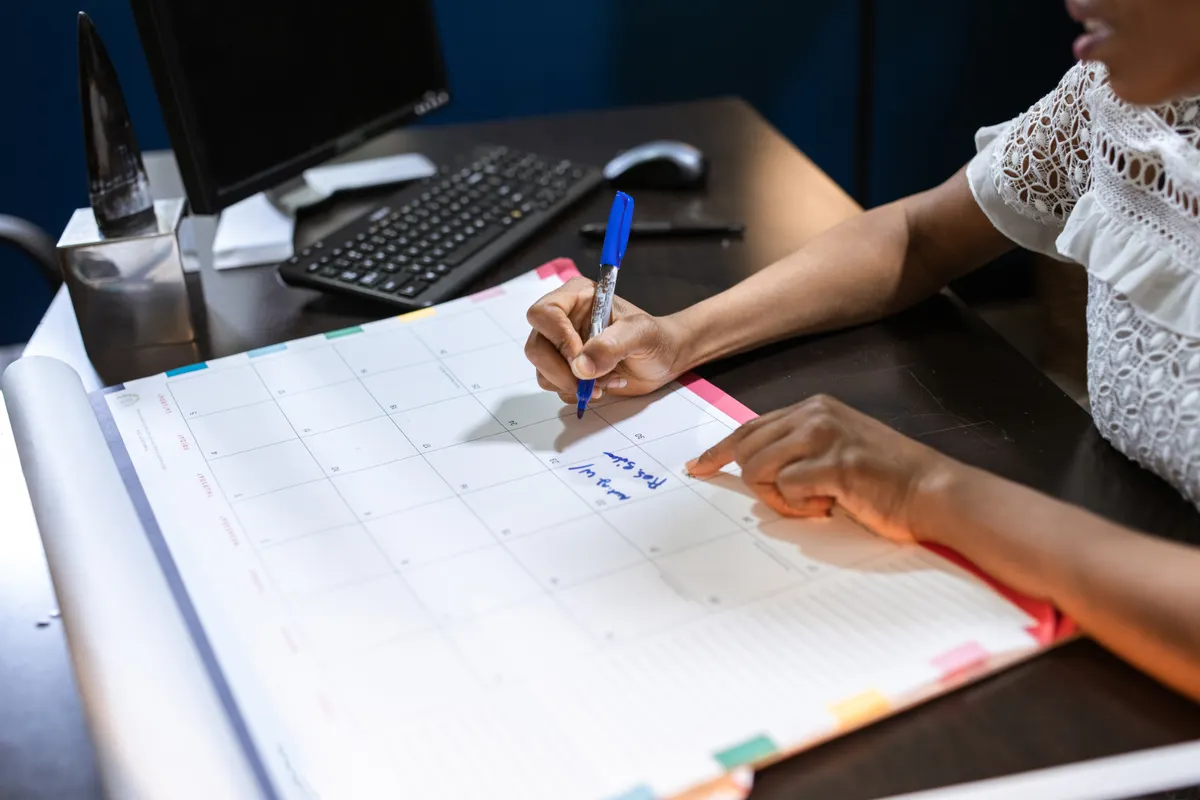 A person at a desk writes on a large paper wall planner beside a computer monitor and keyboard, marking a date with a blue pen.