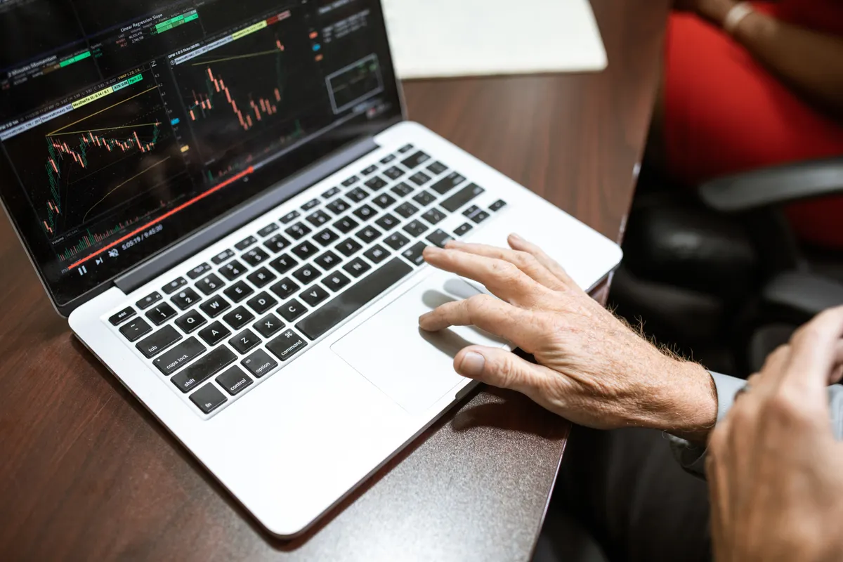 A hand rests on the trackpad of a silver laptop showing a dark chart with candlesticks and trend lines.
