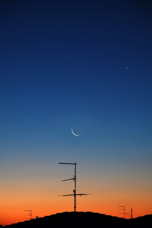 Crescent Moon and Silhouette of an Antenna on a Roof Under Blue Sky