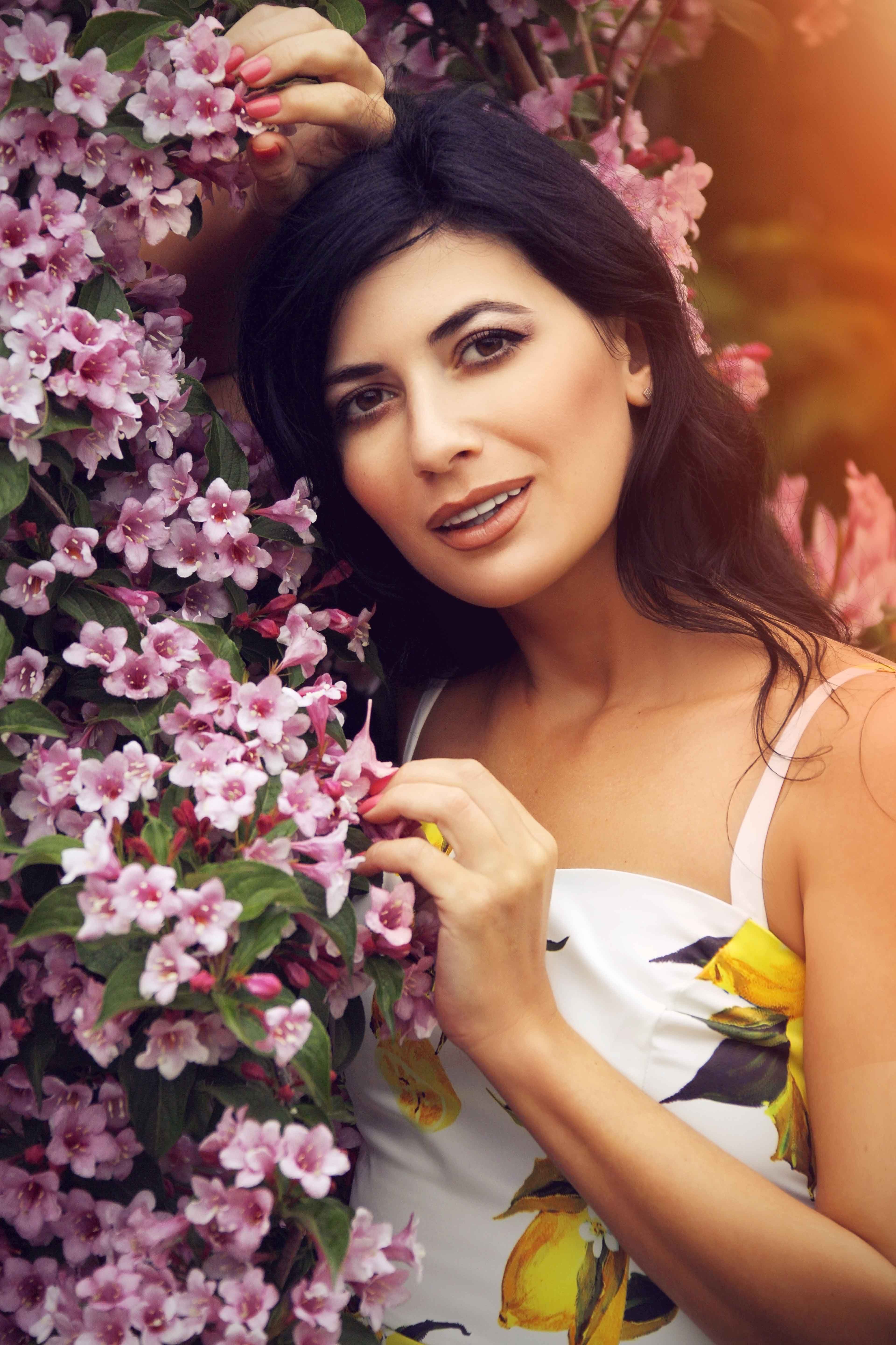 Positive female with long dark hair in casual clothes standing near blooming bush with pink flowers in park and looking at camera
