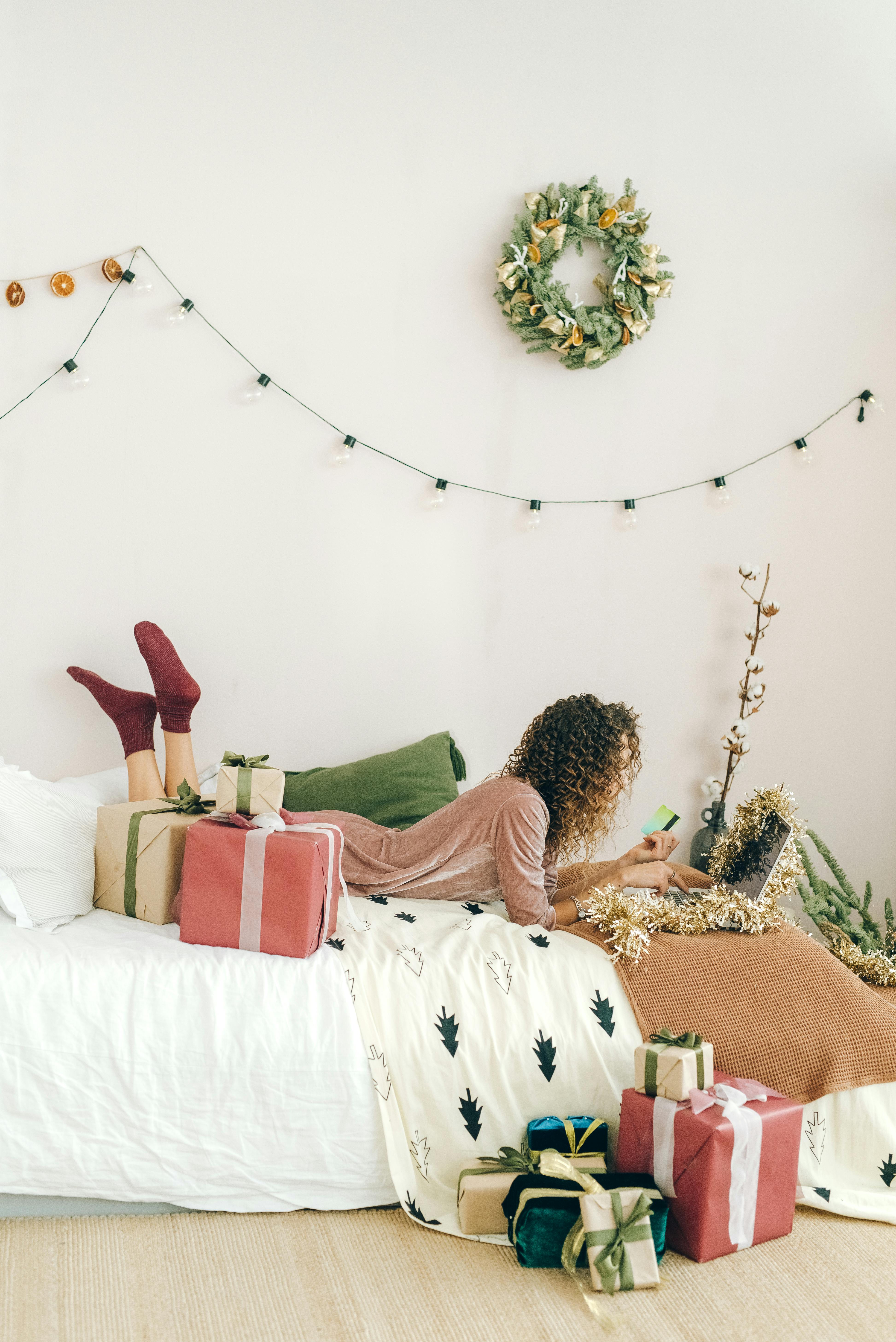 Woman shopping online surrounded by Christmas gifts and decorations in a cozy room.