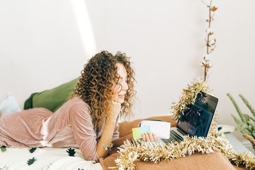 A woman with curly hair shops online using a laptop decorated with Christmas tinsel.