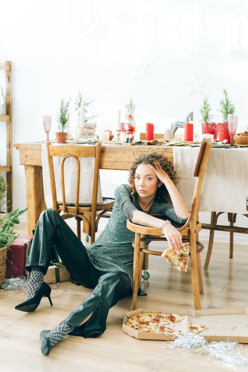 Free A woman in green attire sits on the floor holding pizza, with a decorative table in the background. Stock Photo