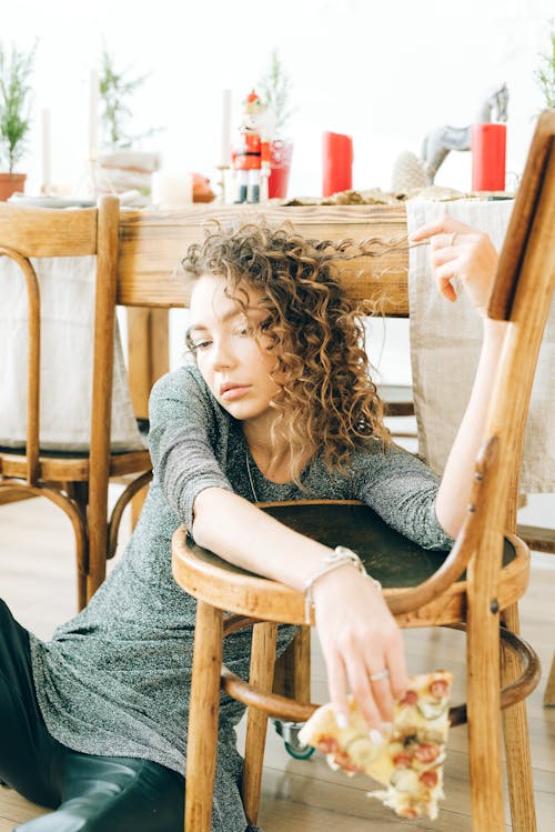 Free Woman Holding a Slice of Pizza Leaning on Brown Wooden Chair Stock Photo